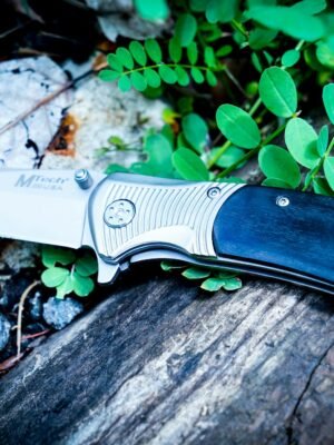 A sharp folding knife with a wooden handle lies on a forest floor surrounded by leaves and rocks.