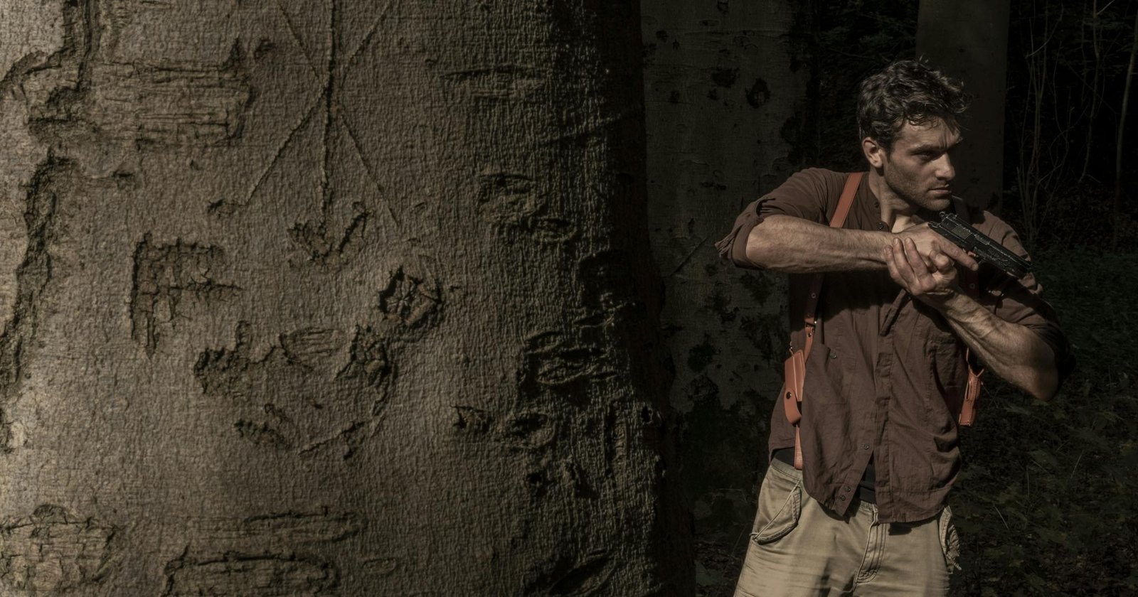 Man camouflaged in forest setting, holding a gun with intent near a tree with carvings.